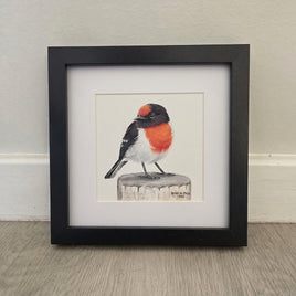 A framed bird art drawing featuring a small bird with a red chest and black-and-white body perched on a log, displayed in a black frame against a light wall on a wood floor.