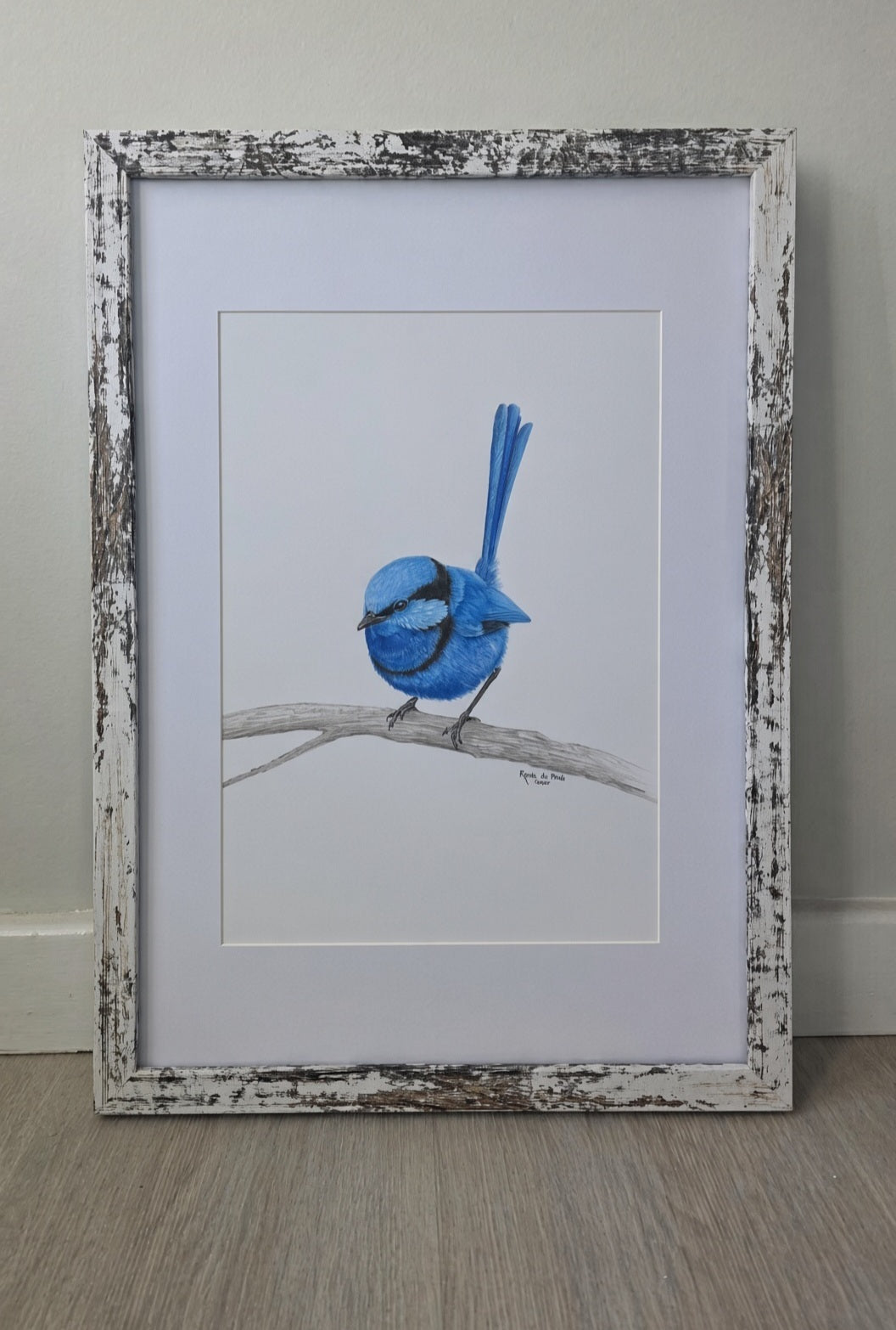 A framed drawing featuring a blue bird perched on a branch, set against a white mat and a distressed white wood frame, leaning against a light gray wall on a wood floor.