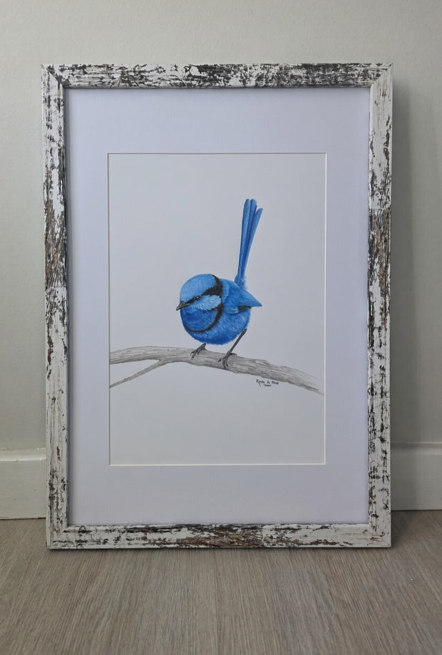 A framed drawing featuring a blue bird perched on a branch, set against a white mat and a distressed white wood frame, leaning against a light gray wall on a wood floor.