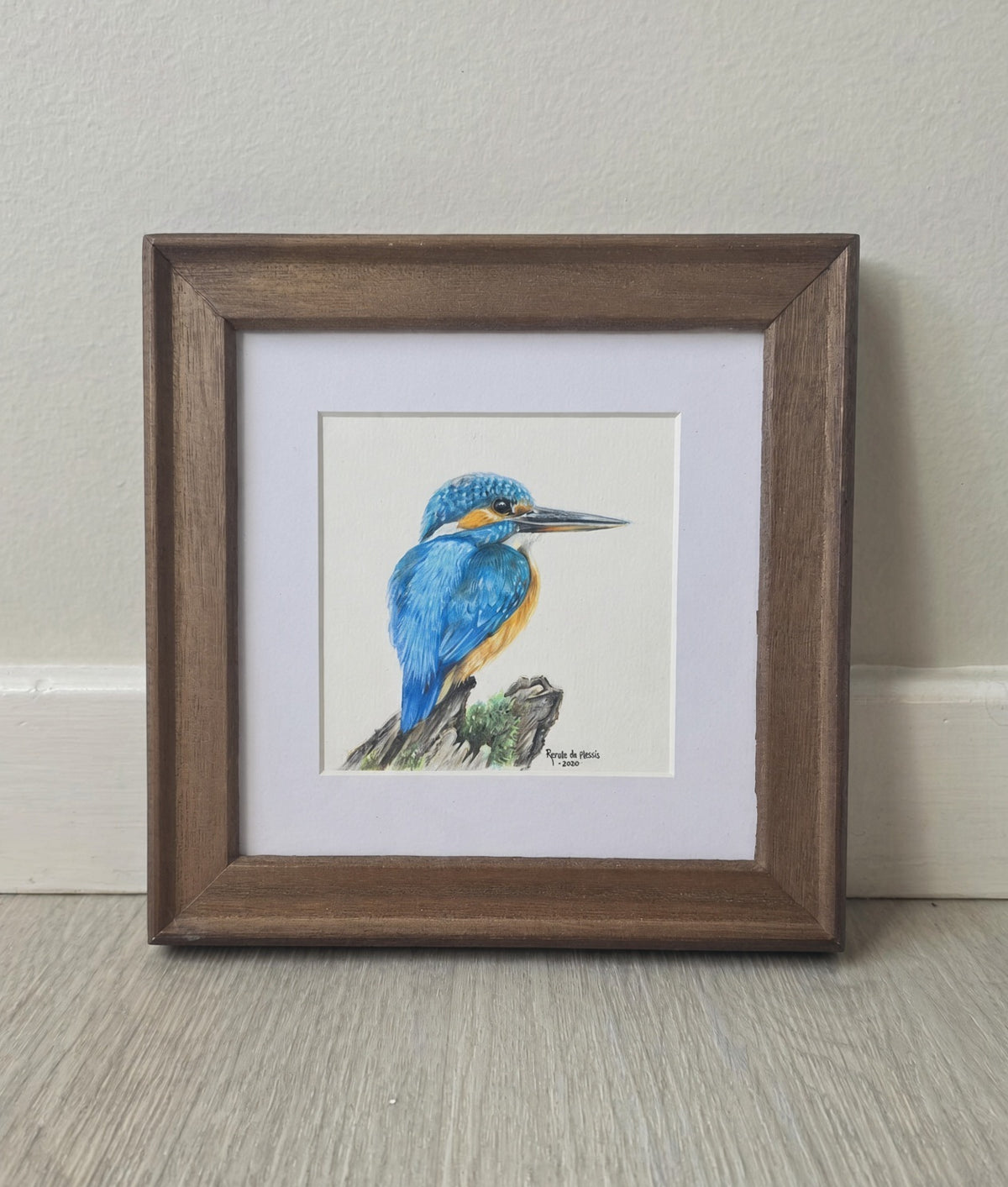 A framed watercolor painting of a blue and orange kingfisher perched on a branch, set within a white mat and a rustic brown wooden frame, leaning against a light beige wall on a gray wood floor.