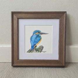 A framed watercolor painting of a blue and orange kingfisher perched on a branch, set within a white mat and a rustic brown wooden frame, leaning against a light beige wall on a gray wood floor.