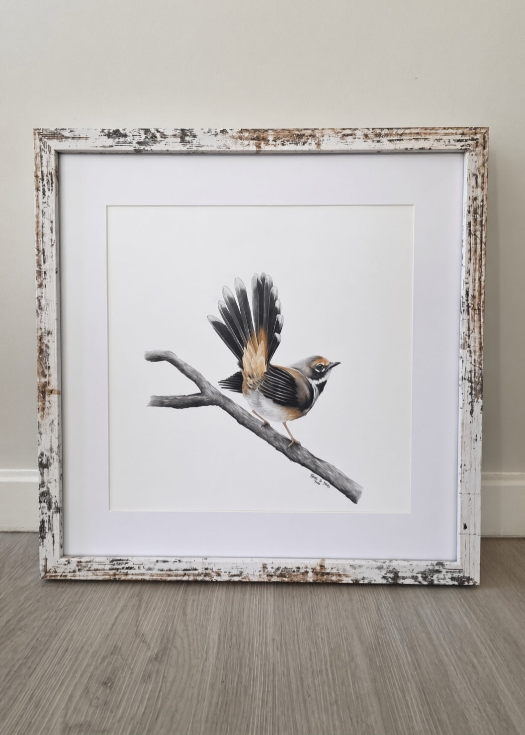 A framed bird drawing featuring a small brown and gray bird perched on a branch, set in a distressed white and beige rustic frame against a neutral wall and wood floor.