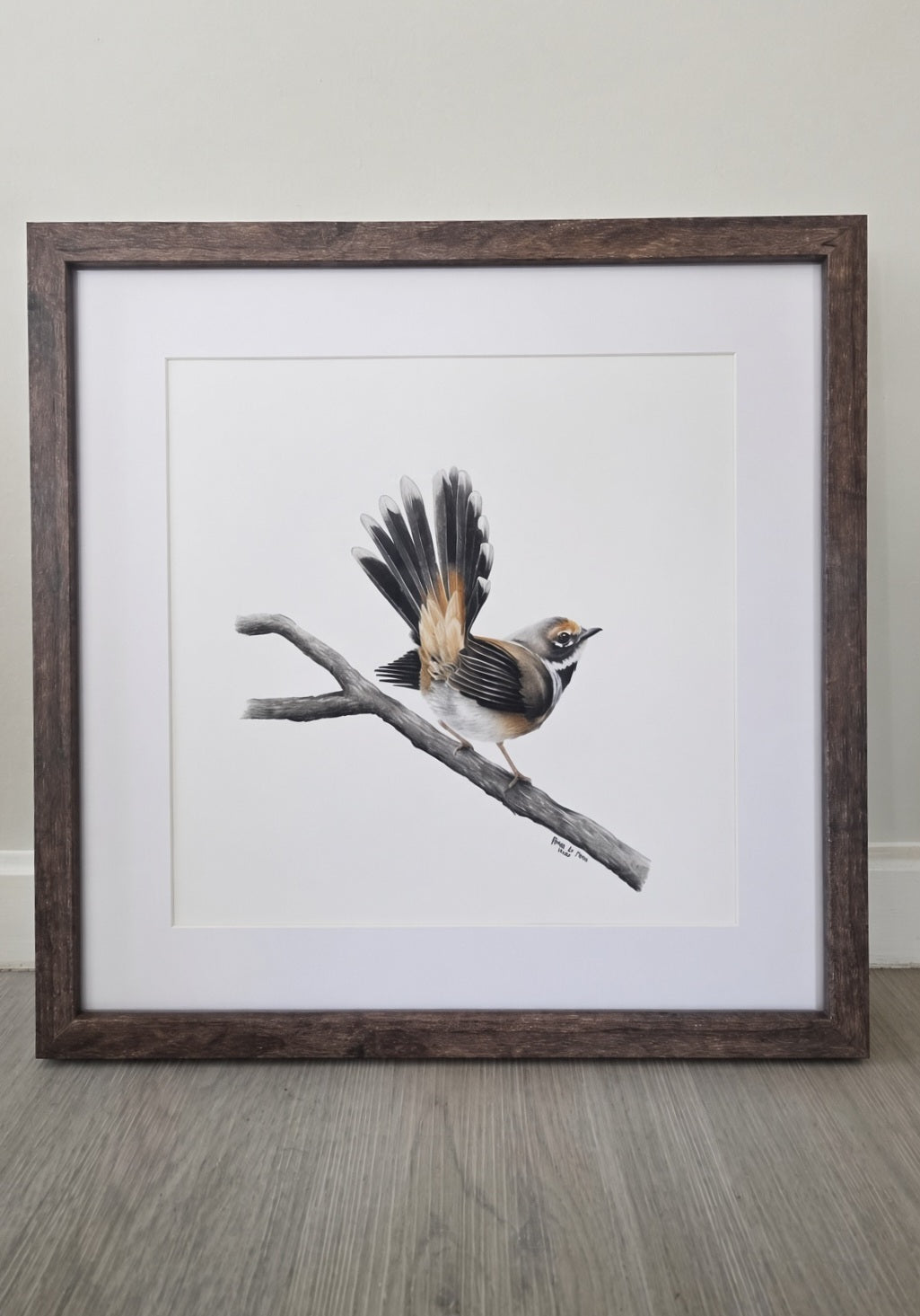 A framed bird art drawing featuring a small brown and white bird perched on a branch, set in a dark wood frame with a white mat against a neutral wall and wood floor.