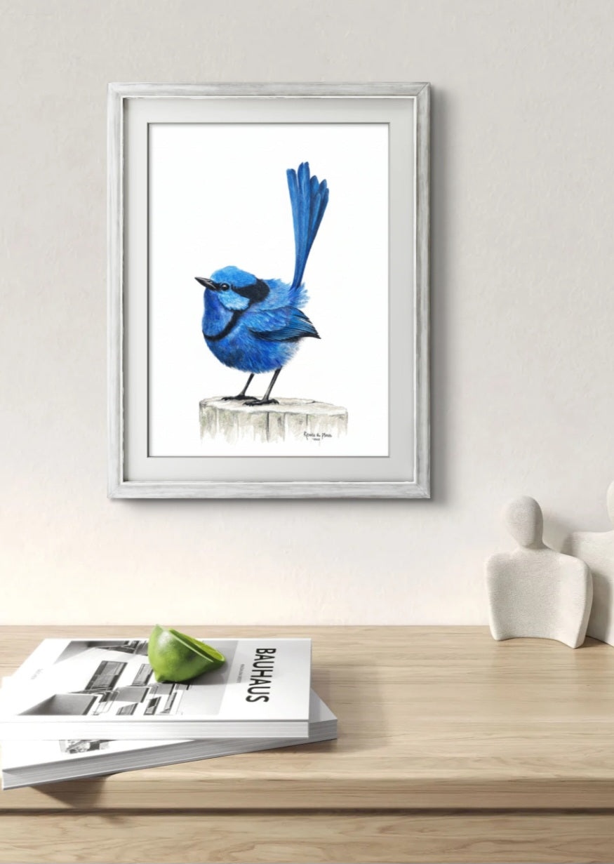 Framed artwork of a blue bird on a white wall above a wooden surface with magazines.