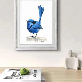 Framed artwork of a blue bird on a white wall above a wooden surface with magazines.