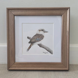 A framed bird illustration drawing showing a kookaburra perched on a branch, set in a white mat and a medium brown wooden frame, resting on a light wood floor against a neutral wall.