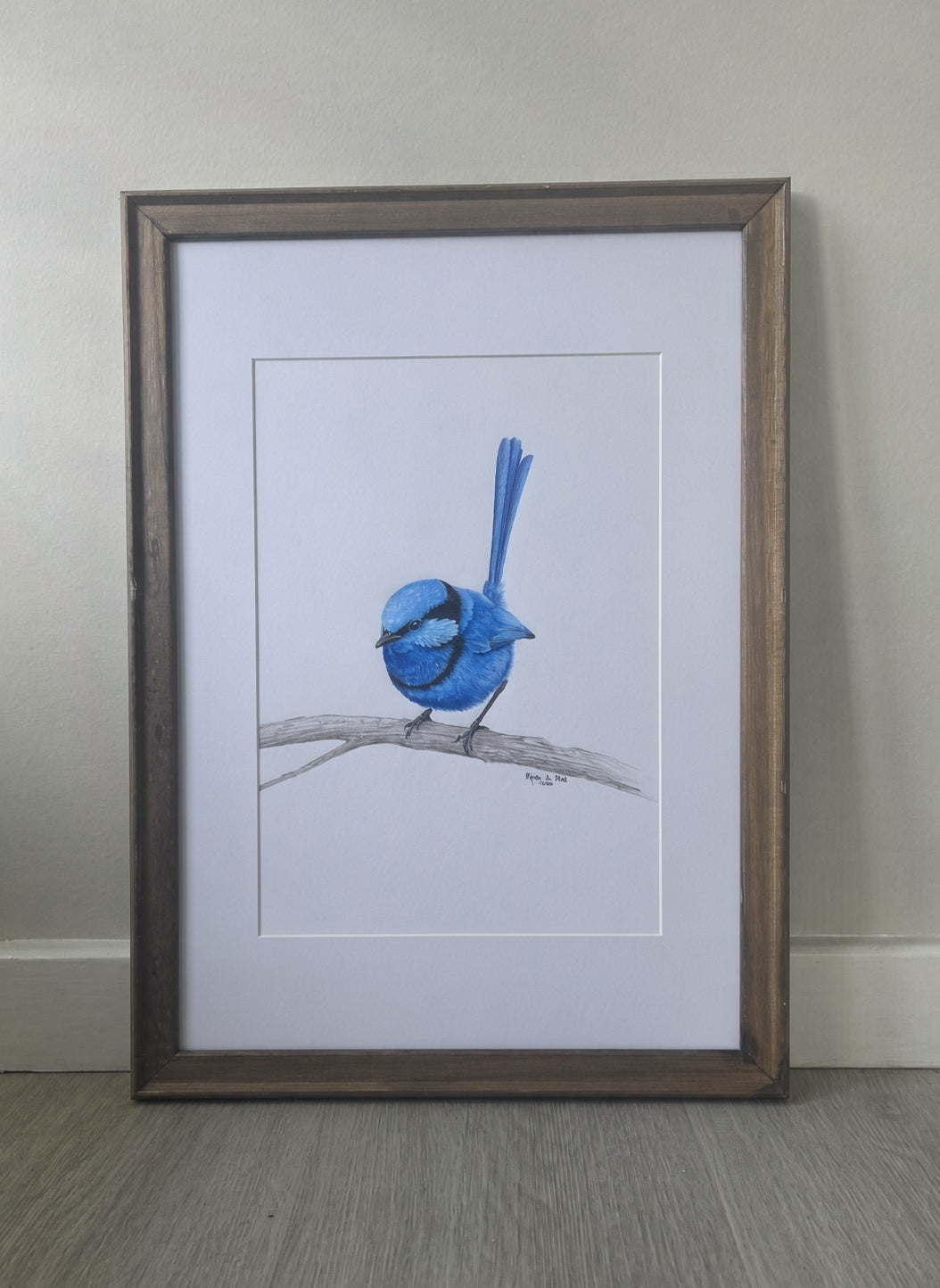 A framed drawing of a blue bird perched on a branch, set against a white mat and a rustic brown wooden frame, leaning against a neutral wall on a wood floor.