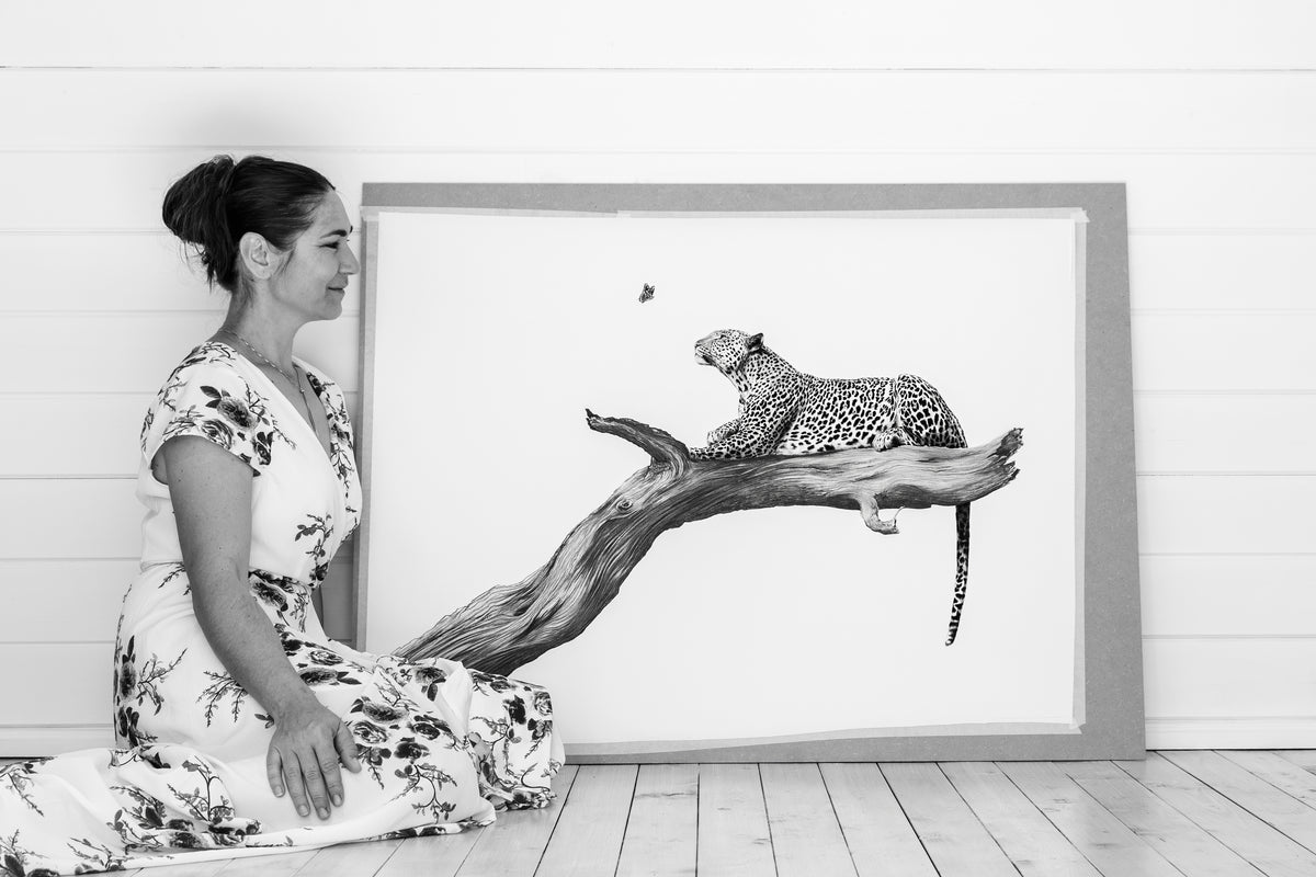 A woman in a floral dress kneels beside a large framed black-and-white artwork showing a leopard resting on a branch against a white paneled wall and floor.