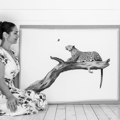 A woman in a floral dress kneels beside a large framed black-and-white artwork showing a leopard resting on a branch against a white paneled wall and floor.