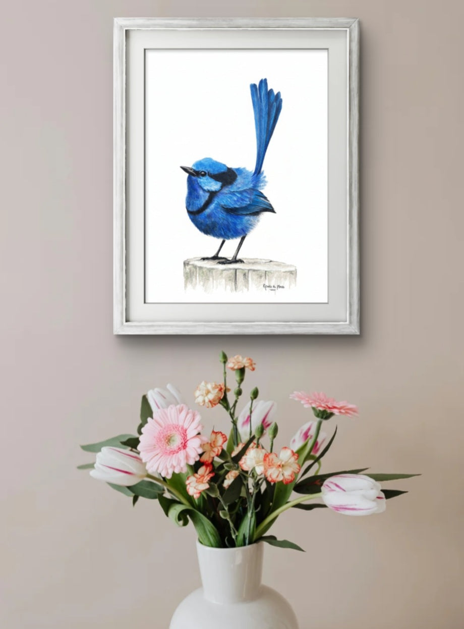 Framed artwork of a blue bird on a white wall with a vase of flowers below.