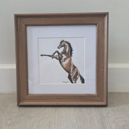 A framed art print featuring a brown horse rearing up, set in a white mat and a medium brown wooden frame, leaning against a light-colored wall on a wood floor.