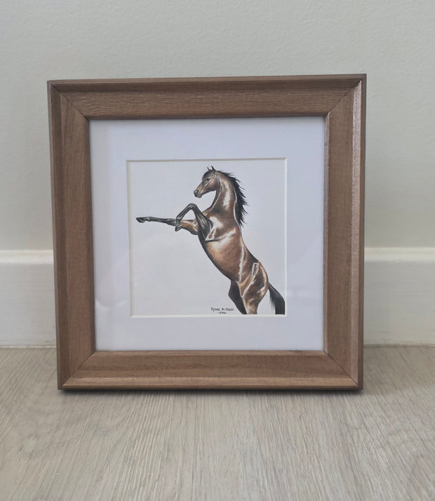 A framed art print featuring a brown horse rearing up, set in a white mat and a medium brown wooden frame, leaning against a light-colored wall on a wood floor.