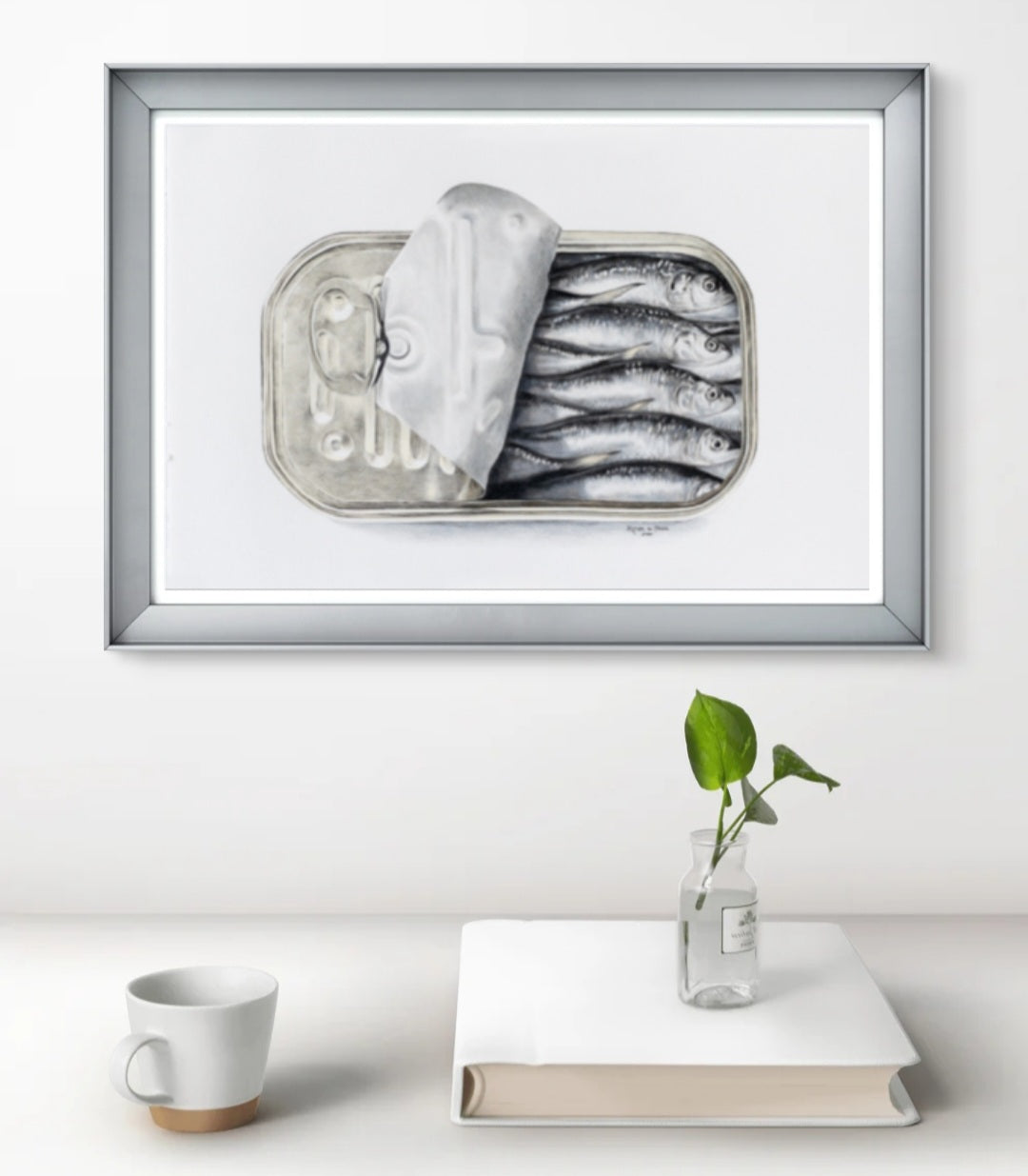 Framed artwork of sardines in a can on a white surface with a plant and book.