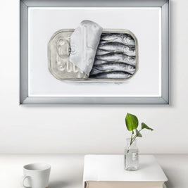 Framed artwork of sardines in a can on a white surface with a plant and book.