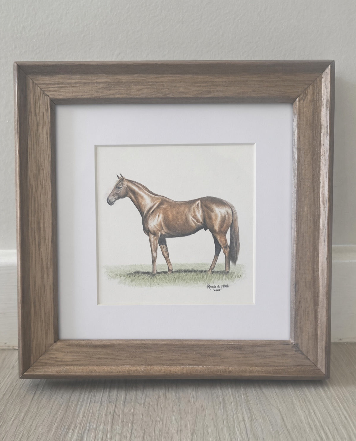 A framed miniature colour pencil drawing featuring a painted brown horse, set in a white mat and a rustic brown wooden frame, resting on a light wood floor against a neutral wall.